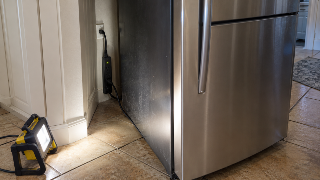 A surge protector with a glowing indicator lamp plugged into a wall receptacle beside a built-in refrigerator pulled slightly forward.